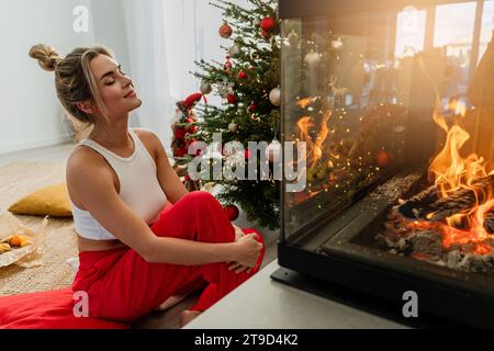 Giovane donna siede accanto a un caminetto luminoso in un accogliente soggiorno adornato con un albero di Natale e decorazioni festive. Foto Stock