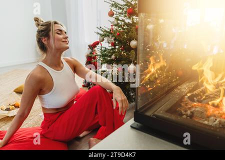 Giovane donna siede accanto a un caminetto luminoso in un accogliente soggiorno adornato con un albero di Natale e decorazioni festive. Foto Stock