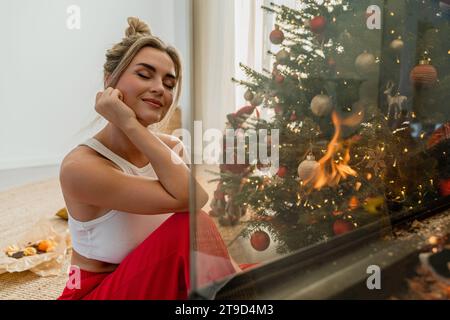 Giovane donna siede accanto a un caminetto luminoso in un accogliente soggiorno adornato con un albero di Natale e decorazioni festive. Foto Stock