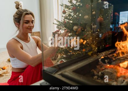 Giovane donna siede accanto a un caminetto luminoso in un accogliente soggiorno adornato con un albero di Natale e decorazioni festive. Foto Stock