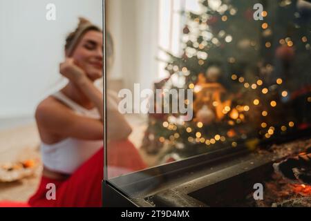 Giovane donna siede accanto a un caminetto luminoso in un accogliente soggiorno adornato con un albero di Natale e decorazioni festive. Foto Stock