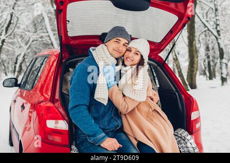 Ritratto di una coppia innamorata che abbraccia il bagagliaio di un'auto nella nevosa foresta invernale. Persone che si rilassano all'aperto durante il viaggio indossando abiti caldi. Foto Stock