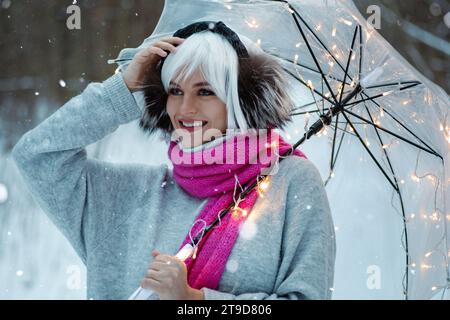 Donna, decorata con parrucca bianca, cuffie termiche, comodo maglione e sciarpa rosa. Ha un ombrello trasparente con luci scintillanti, creato Foto Stock