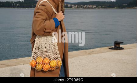 Ragazza con cappotto beige che tiene in mano mandarini arancioni brillanti in una borsa con corda beige. Foto Stock