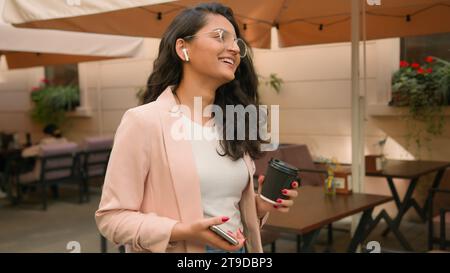 Moderna donna etnica araba indiana sorridente di successo studentessa donna d'affari che cammina in strada caffè della città parlare parlare parlare telefonare cellulare Foto Stock