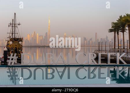 Dubai, Emirati Arabi Uniti, 07.02.22. Insegna Dubai Creek Harbour vicino alla piscina d'acqua turchese, alla nave Pirata Black Pearl e allo skyline del centro di Dubai nei colori dorati dell'alba Foto Stock