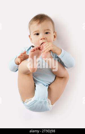 Adorabile bambino in un romper sta mentendo e mette il piede in bocca su sfondo bianco. Foto Stock