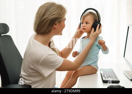 La giovane madre freelance indossa le cuffie wireless con il figlio piccolo seduto alla scrivania del suo ufficio domestico. Foto Stock