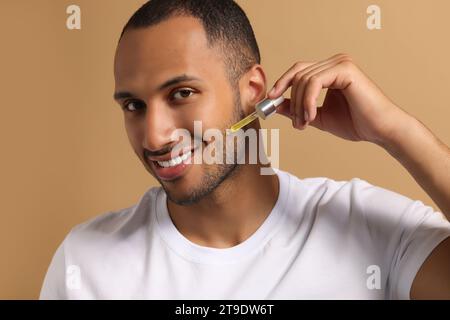 Bell'uomo che applica siero cosmetico sul viso su sfondo marrone chiaro Foto Stock