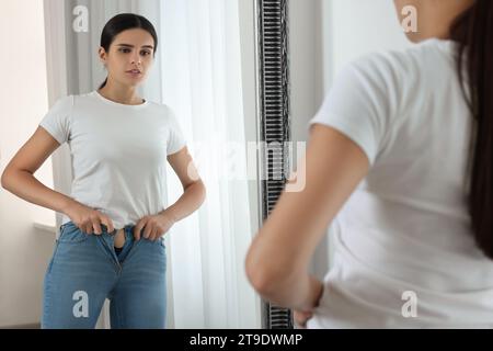 Giovane donna che cerca di indossare jeans stretti vicino allo specchio in casa Foto Stock