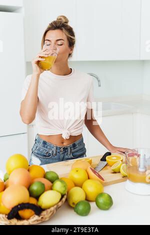 Giovane donna che beve succo d'arancia fatto in casa appena spremuto in cucina bianca Foto Stock