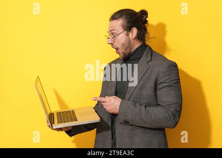 Un uomo barbuto sorpreso che indossa occhiali che puntano sullo schermo del notebook su sfondo giallo Foto Stock