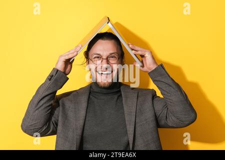 Giovane uomo dalla barba allegro che tiene un computer portatile sopra la testa su sfondo giallo Foto Stock