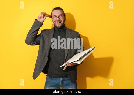 Uomo dalla barba allegra che indossa occhiali con una matita grande e un notebook su sfondo giallo Foto Stock