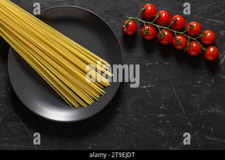 Spaghetti di pasta cruda su piatto grigio e ciuffo di pomodori ciliegini vista dall'alto sfondo scuro, con spazio per copiare il testo. Foto Stock