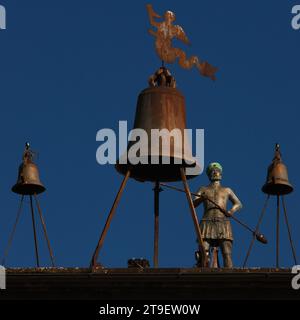 Una figura di bronzo automatizzata colpisce le ore su una grande campana che è il doppio dell'altezza dell'attaccante. Sulla Torre medievale Maurizio (Torre di Maurizio) in Piazza del Duomo di Orvieto, Umbria, Italia. L'orologio meccanico, completato nel 1348, fu commissionato dall'Opera del Duomo (officina del Duomo di Orvieto) per controllare i lavoratori del cantiere della cattedrale. Il nome originale dell’orologio era «ariologium de muriccio», ossia l’orologio del cantiere. Il campanello di bronzo raffigura un ufficiale dell'Opera de Duomo, una Dottiere. Foto Stock