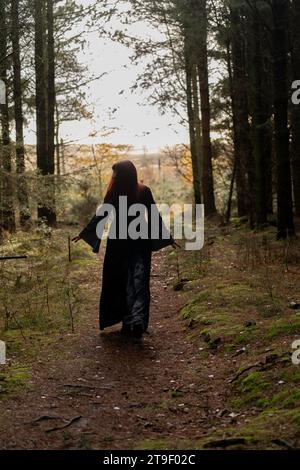 donna in abito nero e grigio con maniche da pipistrello e teschi che cammina verso la macchina fotografica in una foresta Foto Stock