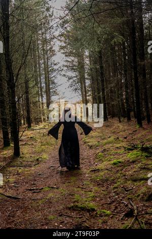 donna in abito nero e grigio con maniche da pipistrello e teschi che cammina verso la macchina fotografica in una foresta Foto Stock