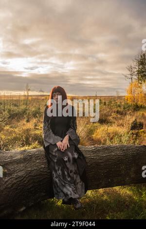 donna in abito nero e grigio con maniche da pipistrello e teschi sedeva su un albero caduto in una foresta Foto Stock