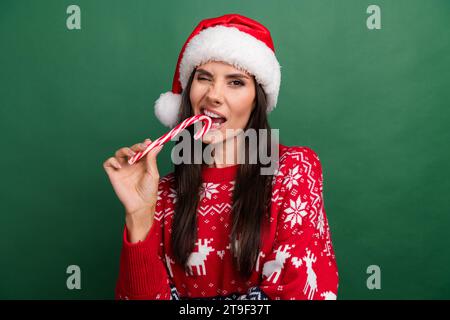 Foto di una bella e funky dama di babbo natale con stampa di natale che morde uno sfondo di colore verde isolato di canna da zucchero Foto Stock