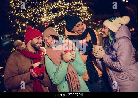 Foto di quattro amici positivi e dolci che indossano i frangivento che si godono le bevande calde e gustose di Natale fuori dal parco del mercato urbano Foto Stock