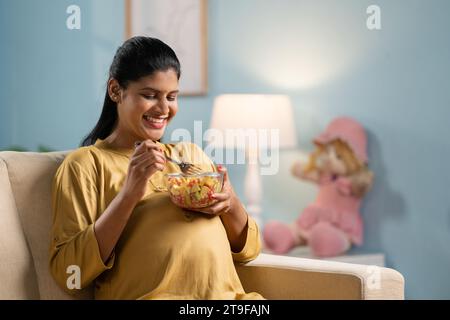 Donna incinta indiana felice che mangia insalata di frutta seduto sul divano a casa - concetto di stili di vita sani, nutrizione materna di gravidanza. Foto Stock