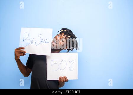 Allegro uomo africano che indossa una camicia nera con una tessera bianca con una grande vendita e un'iscrizione al 50%, annunciando le grandi vendite del Black Friday. isolato su bac blu Foto Stock