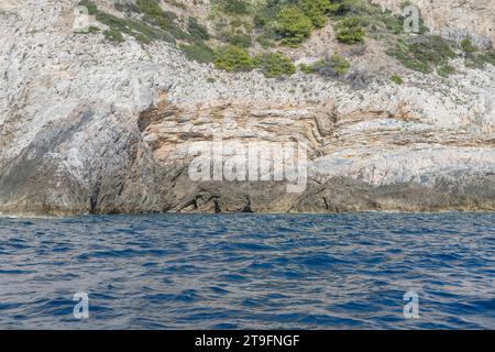 Dettaglio di rocce ignee stratificate su ripide scogliere stratificate di capo piatti al promontorio sulla riva mediterranea, scattate da una barca in luminoso inizio autunno Foto Stock