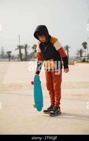 Ritratto verticale a tutta lunghezza di un adolescente multietnico, elegante ragazzo in abbigliamento sportivo, con skateboard, in piedi su un playgr per skatepark all'aperto Foto Stock