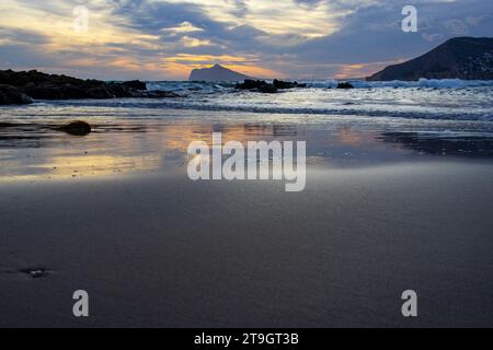 Il tramonto sulla spiaggia si affaccia sulle onde, sulle montagne e sulla sabbia bagnata su cui si riflette il cielo Foto Stock