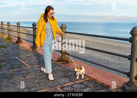 Donna vestita di impermeabile giallo che fa una passeggiata lungo il lungomare con il suo grazioso piccolo Jack Russell Terrier in un pomeriggio primaverile o autunnale. Foto Stock