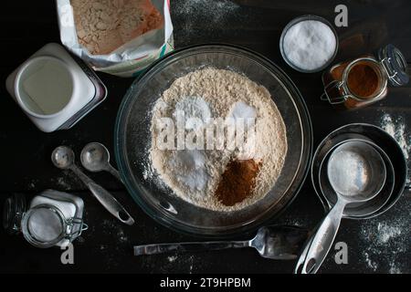 Miscelazione degli ingredienti secchi per le ciambelle di sidro di mele al forno: Farina di pasta integrale in un recipiente di miscelazione in vetro circondato da utensili e ingredienti da cucina Foto Stock