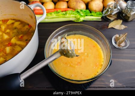 Primo piano di un frullatore a immersione con zuppa di patate sullo sfondo: Frullatore a bastone utilizzato per miscelare la zuppa di patate tedesca (kartoffelsuppe) in un recipiente di vetro Foto Stock