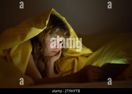 Ragazzo da solo a casa. Ragazzo che guarda un tablet prima di dormire. Il bambino legge l'e-book. Il bambino guarda lo schermo del tablet in alto. Ragazzo che gioca Foto Stock