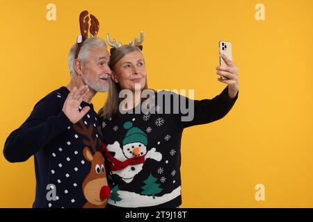 Coppia anziana con maglioni natalizi e fasce di renna che scattano selfie su sfondo arancione. Spazio per il testo Foto Stock
