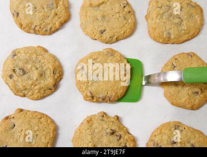 File di biscotti appena sfornati con scaglie di cioccolato e farinata d'avena su carta pergamena, spatola verde in silicone che solleva un biscotto dalla teglia da forno. Foto Stock