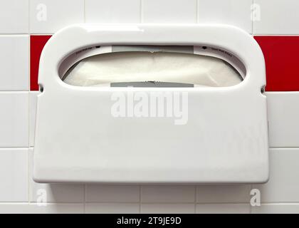 Dispenser di carta igienica coprisedile su parete piastrellata rossa e bianca. Forniture igieniche per il bagno pubblico. Foto Stock