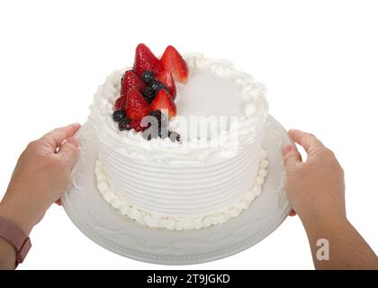 Mani femminili caucasiche mature che tengono in mano una torta alla vaniglia glassata con crema di burro bianca, ricoperta da fragole fresche, boisenberretti e mirtilli Foto Stock