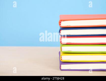 Libri rilegati colorati e luminosi impilati su un tavolo di legno chiaro con sfondo blu. Foto Stock