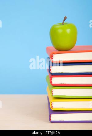 Libri rilegati colorati e luminosi impilati su un tavolo di legno chiaro con sfondo blu. Mela verde sopra i libri impilati. Foto Stock