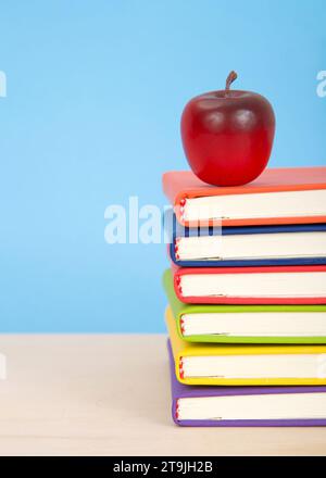 Libri rilegati colorati e luminosi impilati su un tavolo di legno chiaro con sfondo blu. Mela rossa sopra i libri impilati. Foto Stock