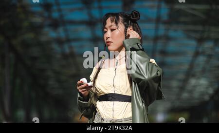 Una bella donna asiatica con pantaloncini corti e un cappotto verde in pelle esce dalla metropolitana e utilizza cuffie wireless. Foto Stock