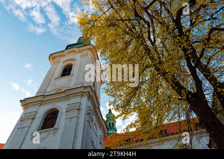 Una chiesa bianca con cupola verde e guglia, circondata da alberi dalle foglie gialle. Albero giallo autunnale vicino al monastero di Strahov a Praga. Foto Stock