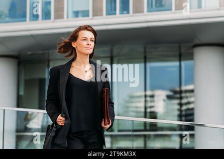 Donna d'affari sicura di mezza età che tiene una cartella in pelle che cammina contro un edificio di uffici Foto Stock