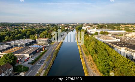 Questa immagine aerea mostra un canale sereno che attraversa un'area urbana, fiancheggiato da un lato da edifici industriali e dall'altro da una lussureggiante vegetazione. Il contrasto tra la calma via d'acqua e la vita urbana adiacente parla della coesistenza di industria e natura entro i confini della città. Sopra, il cielo azzurro e cristallino suggerisce una giornata piena di possibilità, mentre la curva dolce del canale aggiunge un senso di calma al vivace ambiente. Tranquillità urbana: Canale fiancheggiato dal verde e dall'industria. Foto di alta qualità Foto Stock