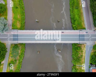 Questa immagine offre una vista aerea di un ponte che attraversa un corpo d'acqua fluente, con auto visibili lungo la sua lunghezza, illustrando il ritmo quotidiano del transito. Il corso d'acqua sottostante, delimitato dal verde, trasporta una corrente dolce e alcune piccole imbarcazioni sono ormeggiate lungo le rive. La simmetria della struttura del ponte contrasta con il flusso organico del fiume, rappresentando l'intersezione tra le realizzazioni realizzate dall'uomo e l'ambiente naturale. Correnti di attraversamento: Vista aerea di un ponte sull'acqua. Foto di alta qualità Foto Stock