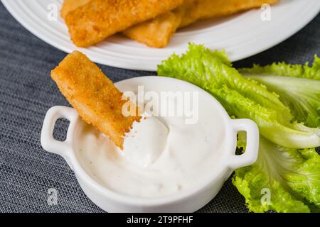 Bastoncini di pesce in una croccante impanatura dorata e una ciotola di salsa di yogurt greco servita con lattuga verde da vicino sul tavolo della cucina Foto Stock