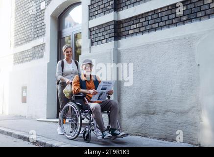 Nipote che spinge un uomo anziano in sedia a rotelle per strada. Donna che si prende cura di sé e anziano che si gode una calda giornata autunnale, tornando a casa dallo shopping. Foto Stock