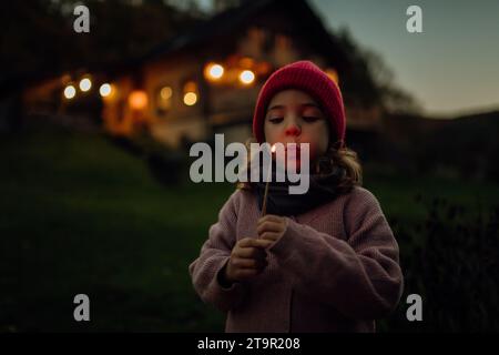 Ritratto di una ragazza carina che soffia la fiamma su un bastoncino in fiamme. La famiglia ha un barbecue in giardino in una sera d'autunno. Foto Stock