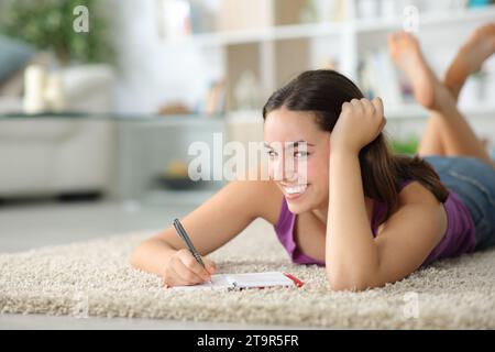 Donna felice sdraiata su un tappeto a casa che scrive all'ordine del giorno guardando te Foto Stock